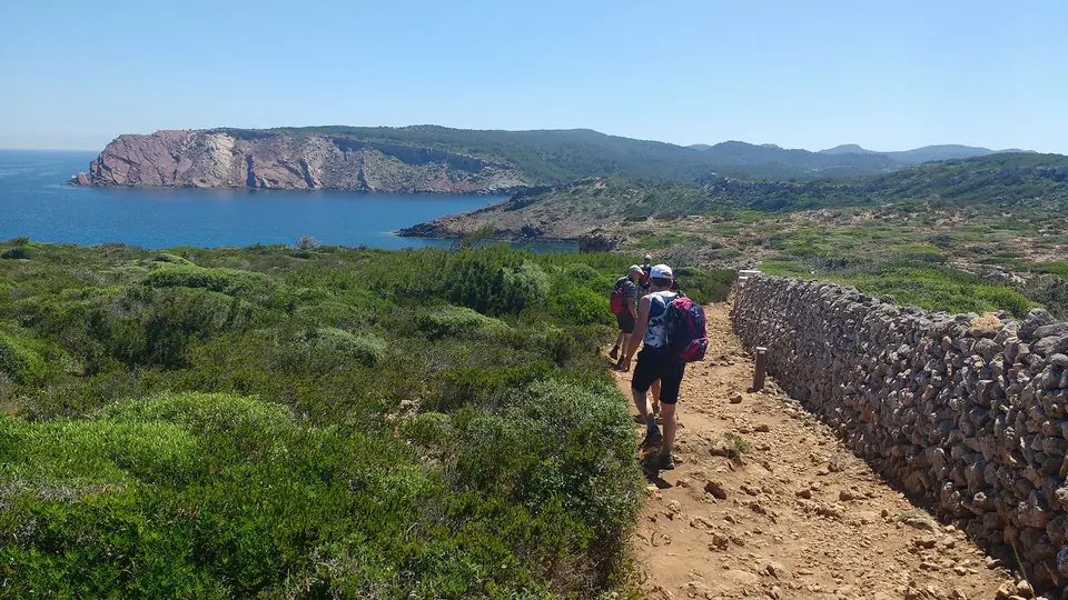 Excursionistes seguint un sender vora una paret de pedra i un cap al fons