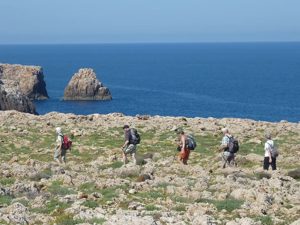 Excursionistes caminant per un terreny de matollar a prop d’uns faralions sobre el mar