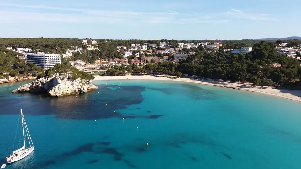 Cala Galdana: badia d’aigües turquesa amb un veler i edificis al fons