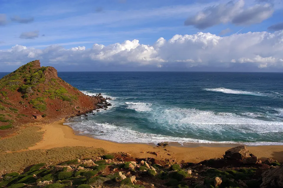 Cala Pilar: platja de sorra fosca amb un penya-segat rogenc i ones