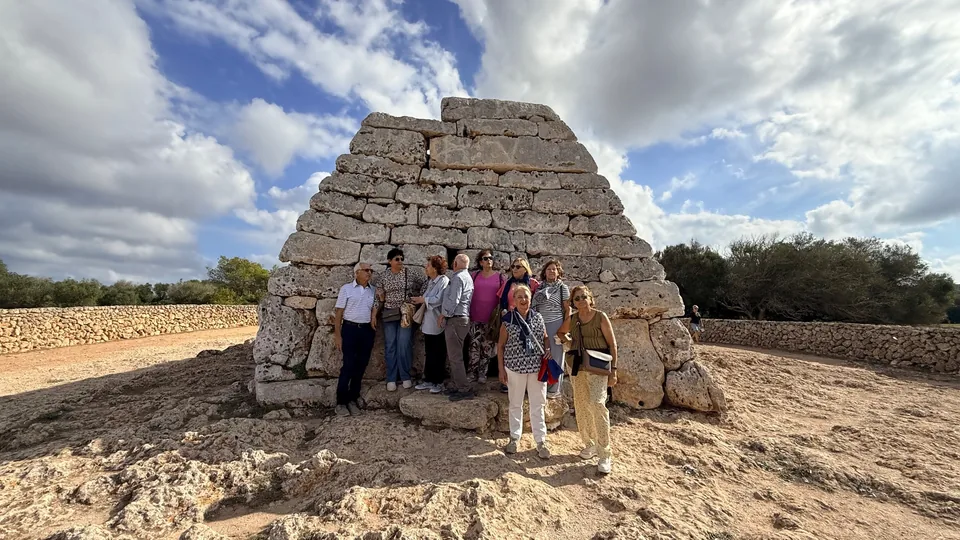 Grup de visitants posant al costat d’una naveta talaiòtica de pedra, amb cel blau i núvols
