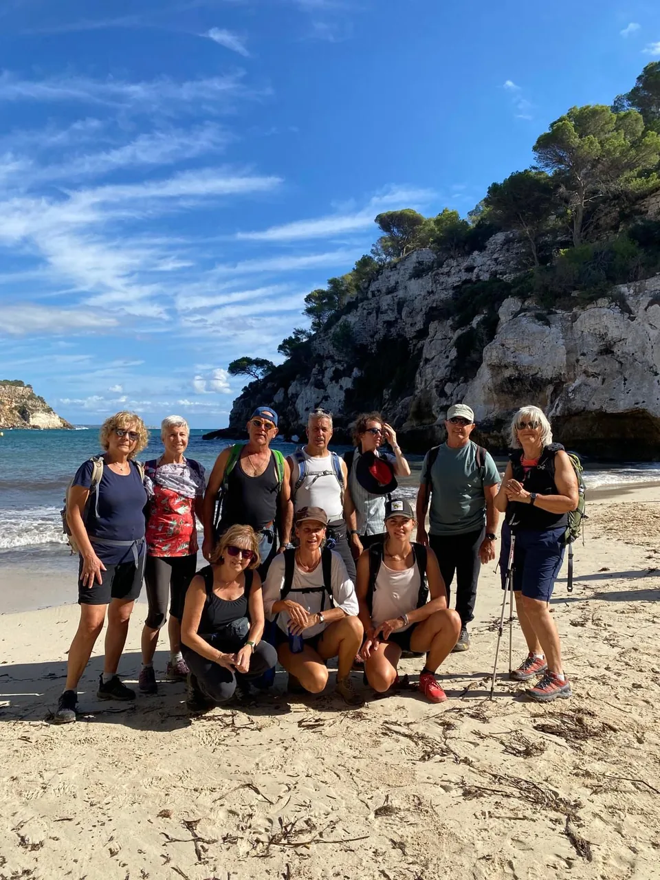 Grup de deu excursionistes posant a la sorra d’una cala, amb un penya-segat i pins al fons